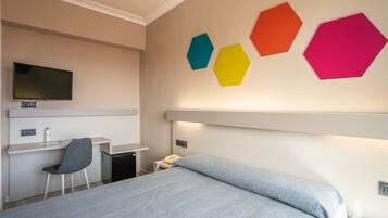 Habitación de hotel con cama, escritorio, silla, televisor montado en la pared y decoración hexagonal de varios colores.