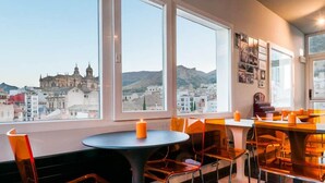 A dining area with round tables and orange chairs, large windows with a cityscape view, and a candle on one table. - Hotel Xauen (Jaén)