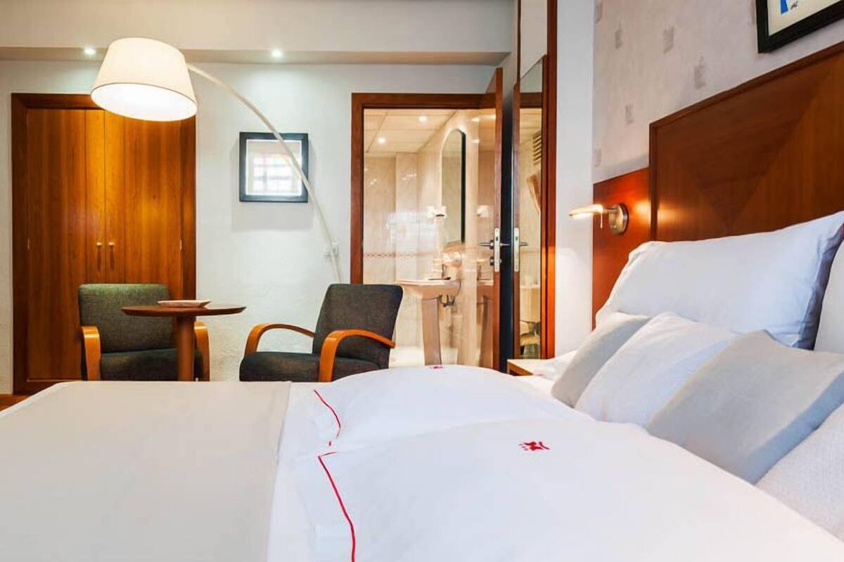 A hotel room with a large bed, a wooden wardrobe, a green armchair, a small round table, and a mirror.