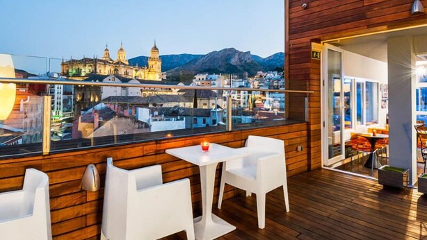 A rooftop terrace with wooden flooring, white chairs, and a table. A cityscape with historic buildings and mountains is visible in the background. - Hotel Xauen (Jaén)