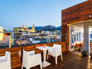 Una terraza en la azotea con suelo de madera, sillas blancas y una mesa. Se aprecia un paisaje urbano con edificios históricos y montañas al fondo.