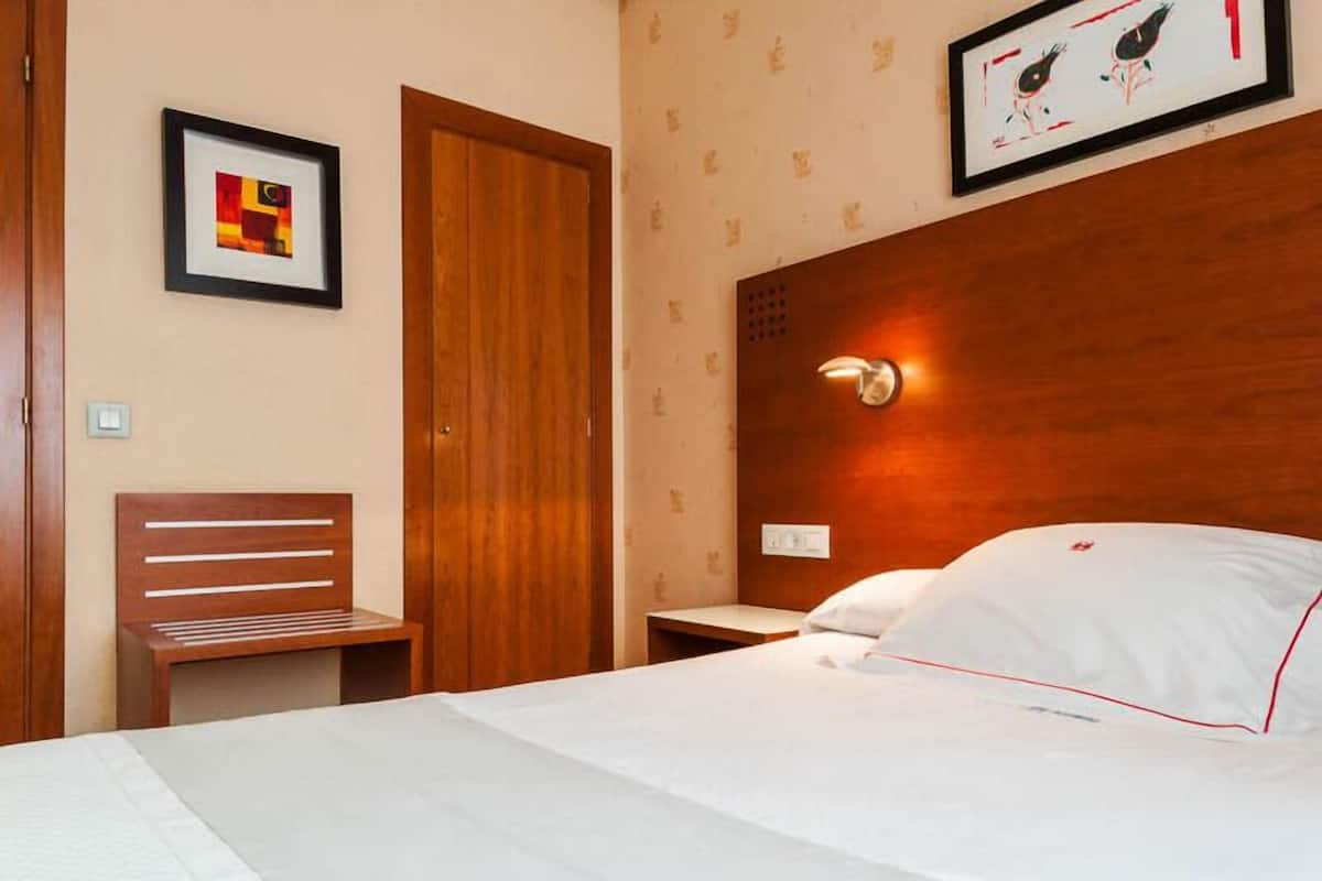 A hotel room with a wooden headboard, a bed with white bedding, a wooden nightstand, a framed picture on the wall, and a wooden door.