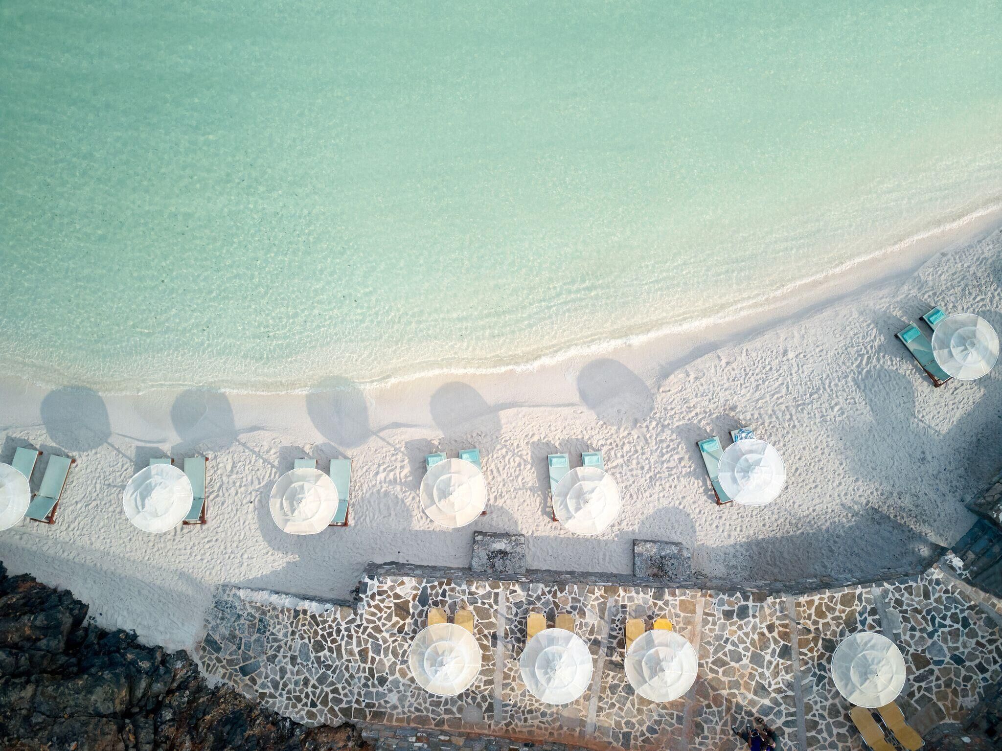 An aerial view of a beach with rows of sun loungers and umbrellas, a stone-paved walkway, and a clear view of the ocean.