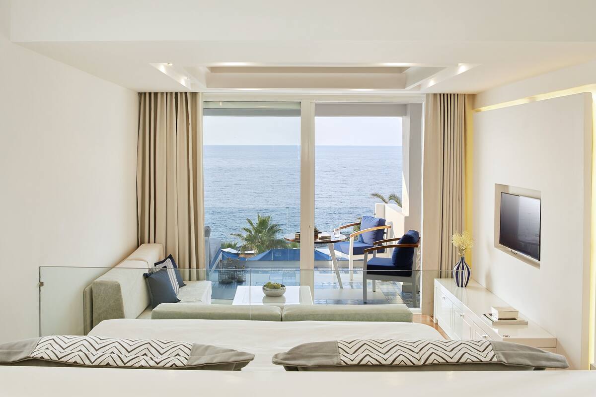 A modern hotel room with a large balcony, a bed, a TV, and a view of the sea.