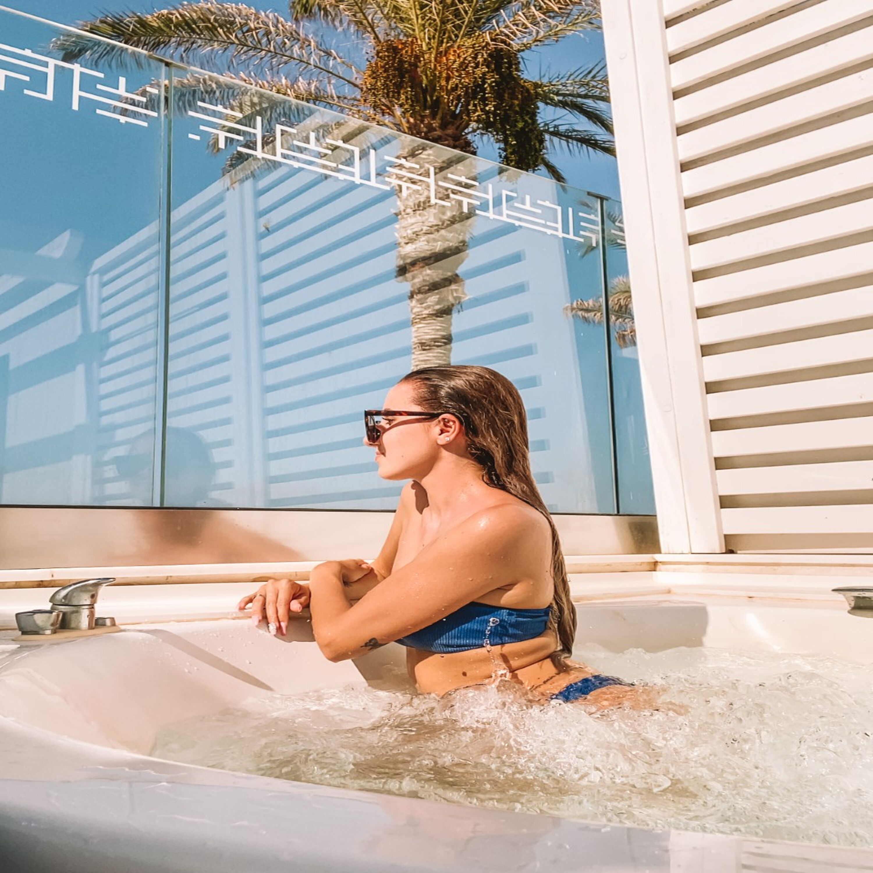 A person in a bathtub with water, a palm tree, and a building reflection.