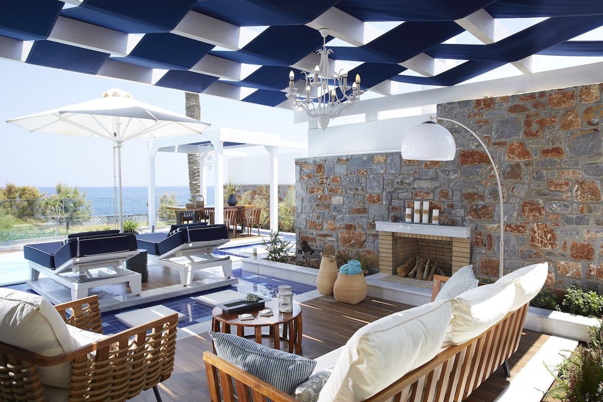A sunlit outdoor lounge area with seating, a stone fireplace, and a view of the sea.