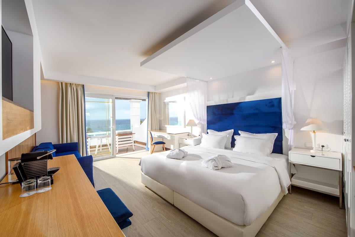 A modern hotel room with a large bed, a desk with a chair, a small table, and a view of the ocean.