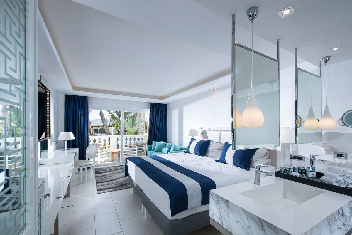 A modern hotel room with a large bed, a separate bathroom with a marble sink, and a balcony with outdoor furniture.