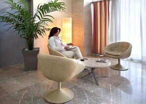 A woman sitting on a chair in a lobby with a potted plant, a lamp, and a table with magazines. - Paolo (Loutraki-Agioi Theodoroi)