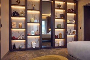 A modern hotel room with a large bed, a built-in wardrobe with illuminated shelves, and a view of the adjacent room.