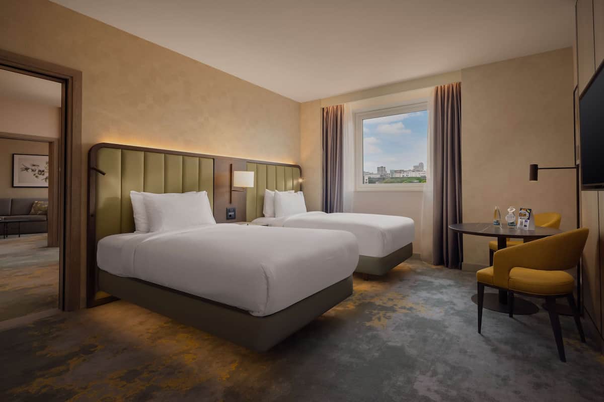 A hotel room with two beds, a small table with chairs, a TV, and a window with a city view.