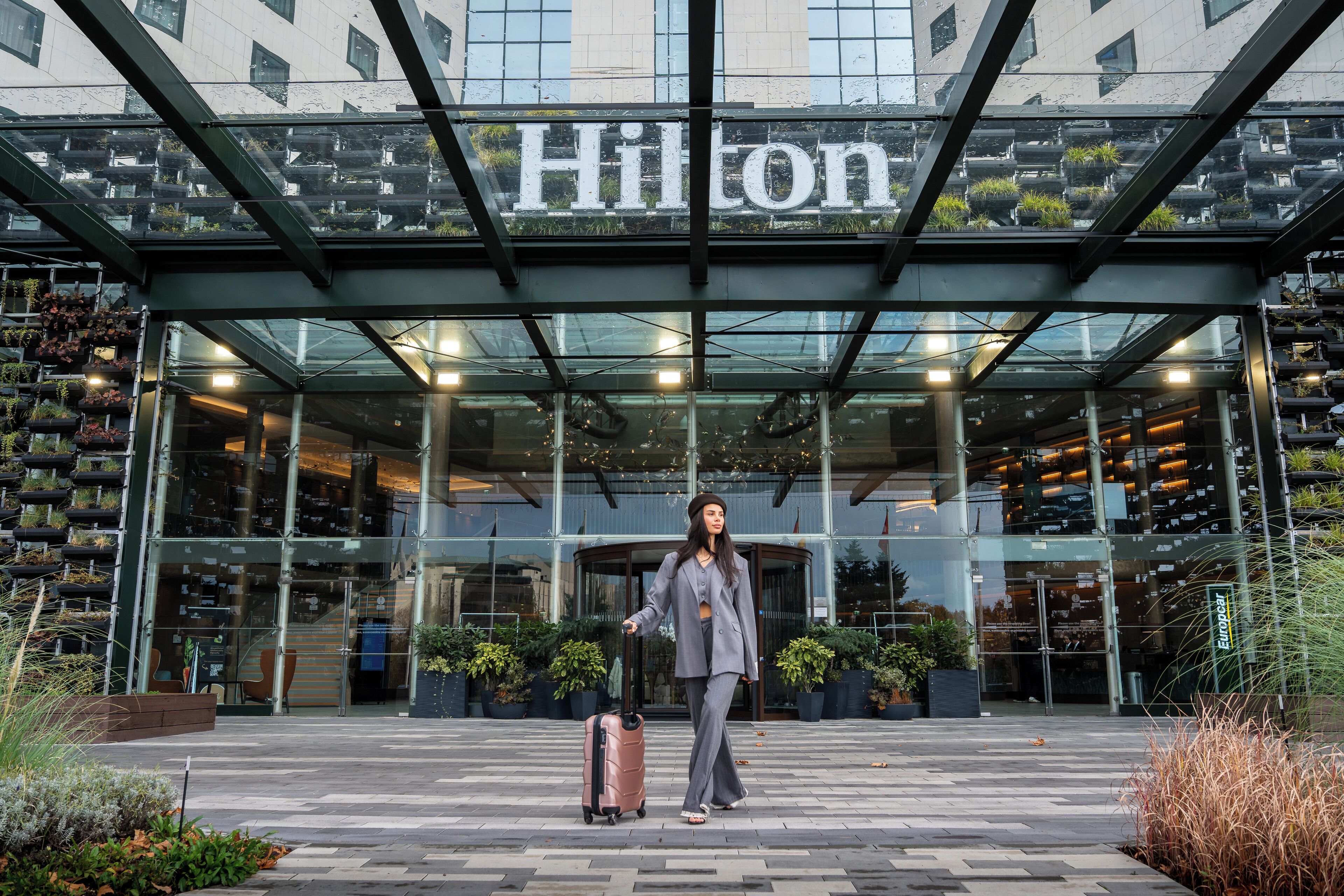 Una entrada de hotel moderna con un toldo de cristal, plantas en macetas y un cartel que dice "Hilton".