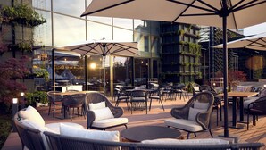 An outdoor seating area with wicker furniture and umbrellas, overlooking a modern building with large windows and greenery.