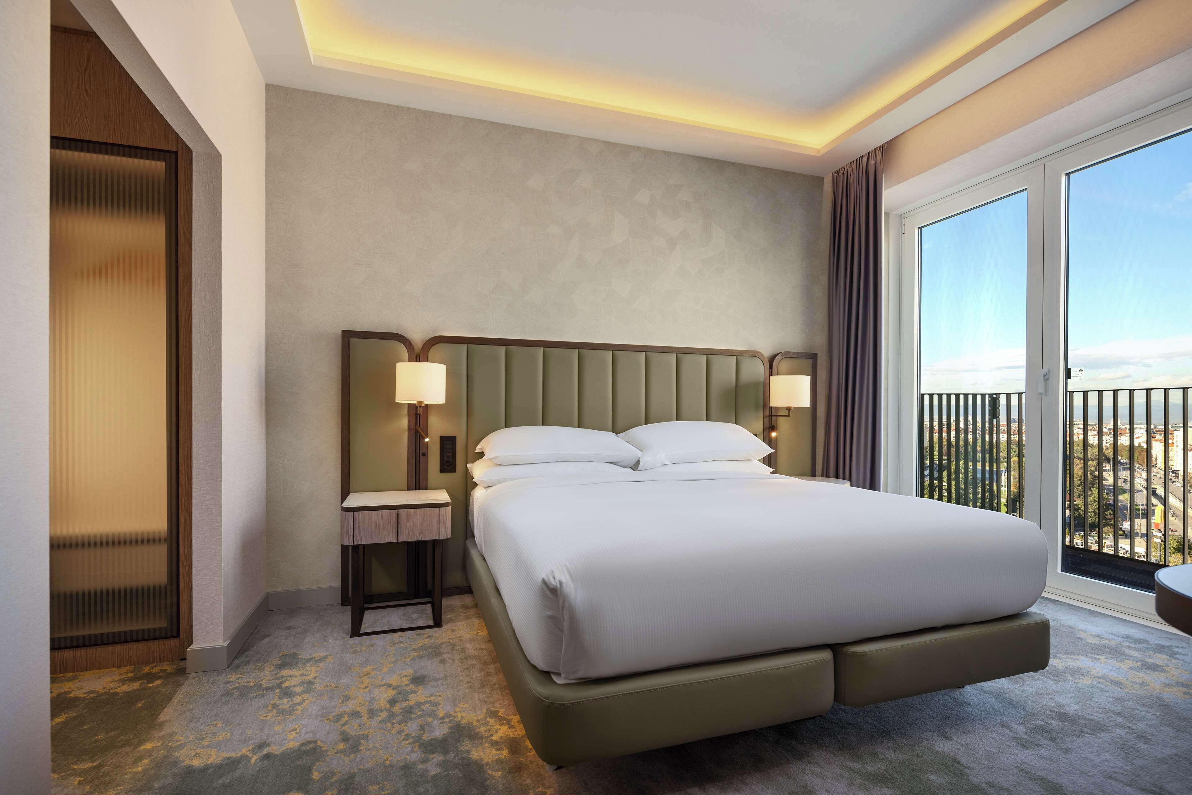 A hotel room with a large bed, two bedside lamps, a nightstand, and a view of the city through large windows.