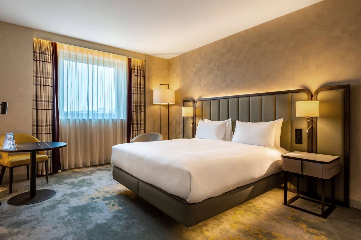 A hotel room with a large bed, bedside lamps, a small table, and a chair.