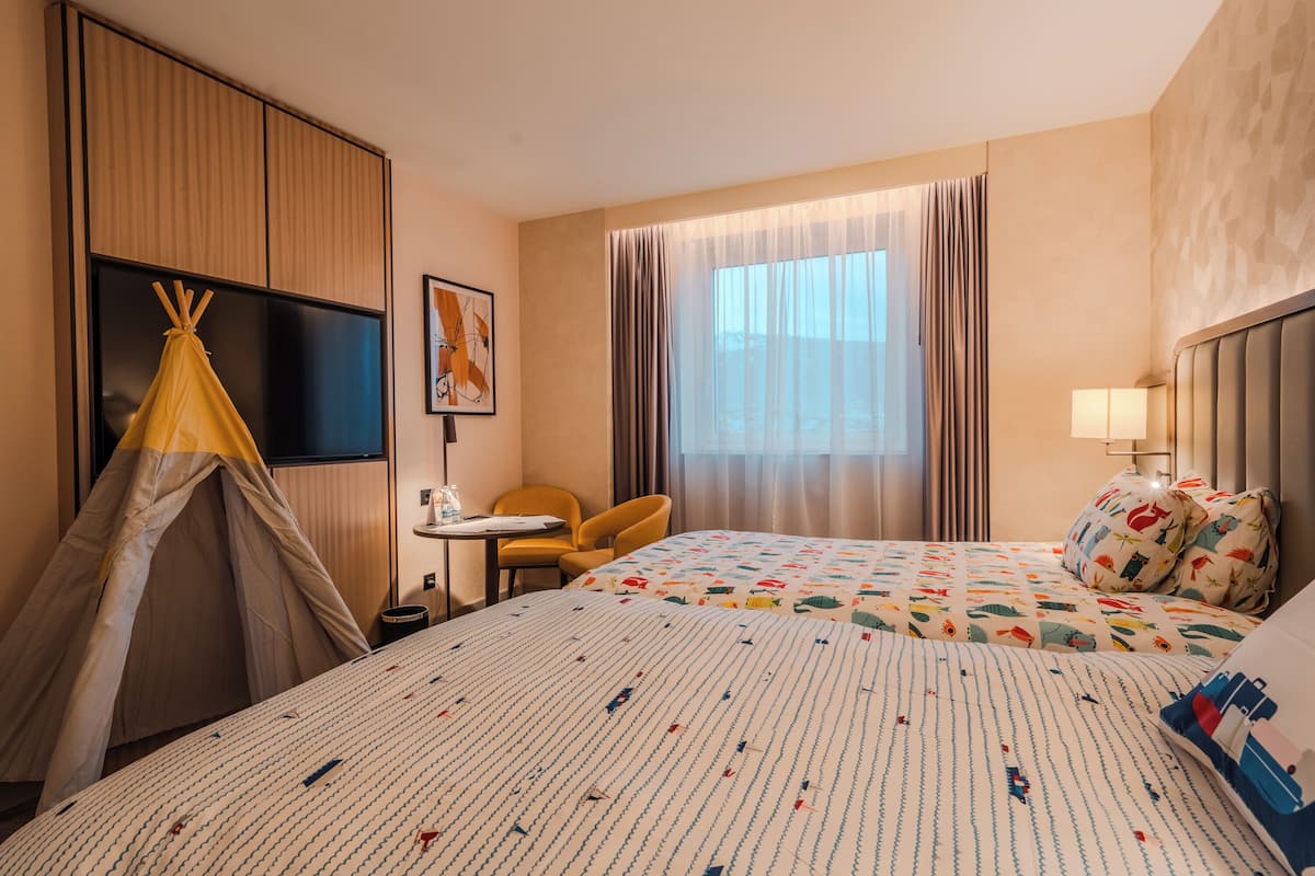 A hotel room with a bed, a chair, a small table, a TV, and a window with curtains.