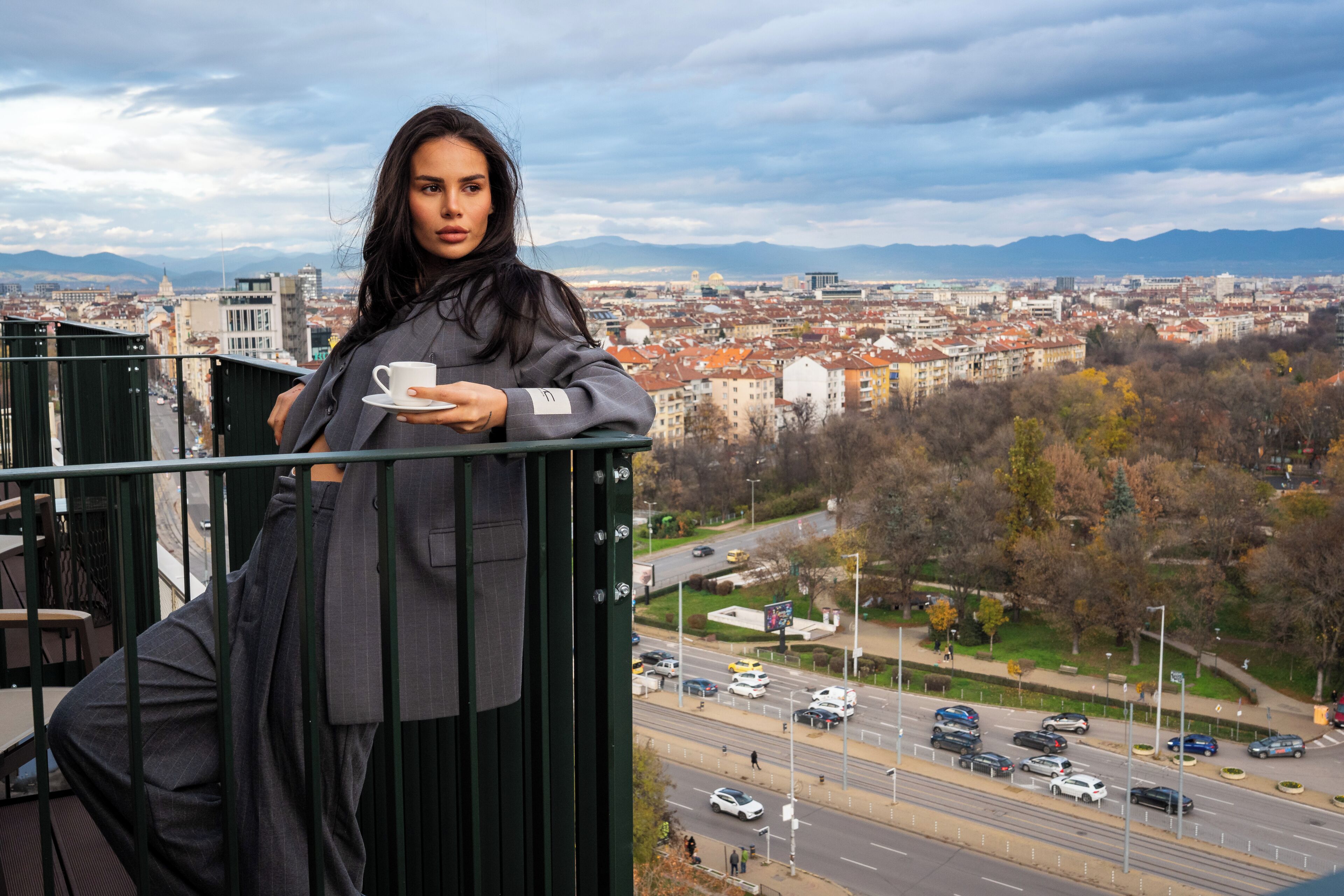 Eine Person hält eine Tasse auf einem Balkon mit Blick auf eine Stadtlandschaft, einschließlich Gebäude und einer Straße.