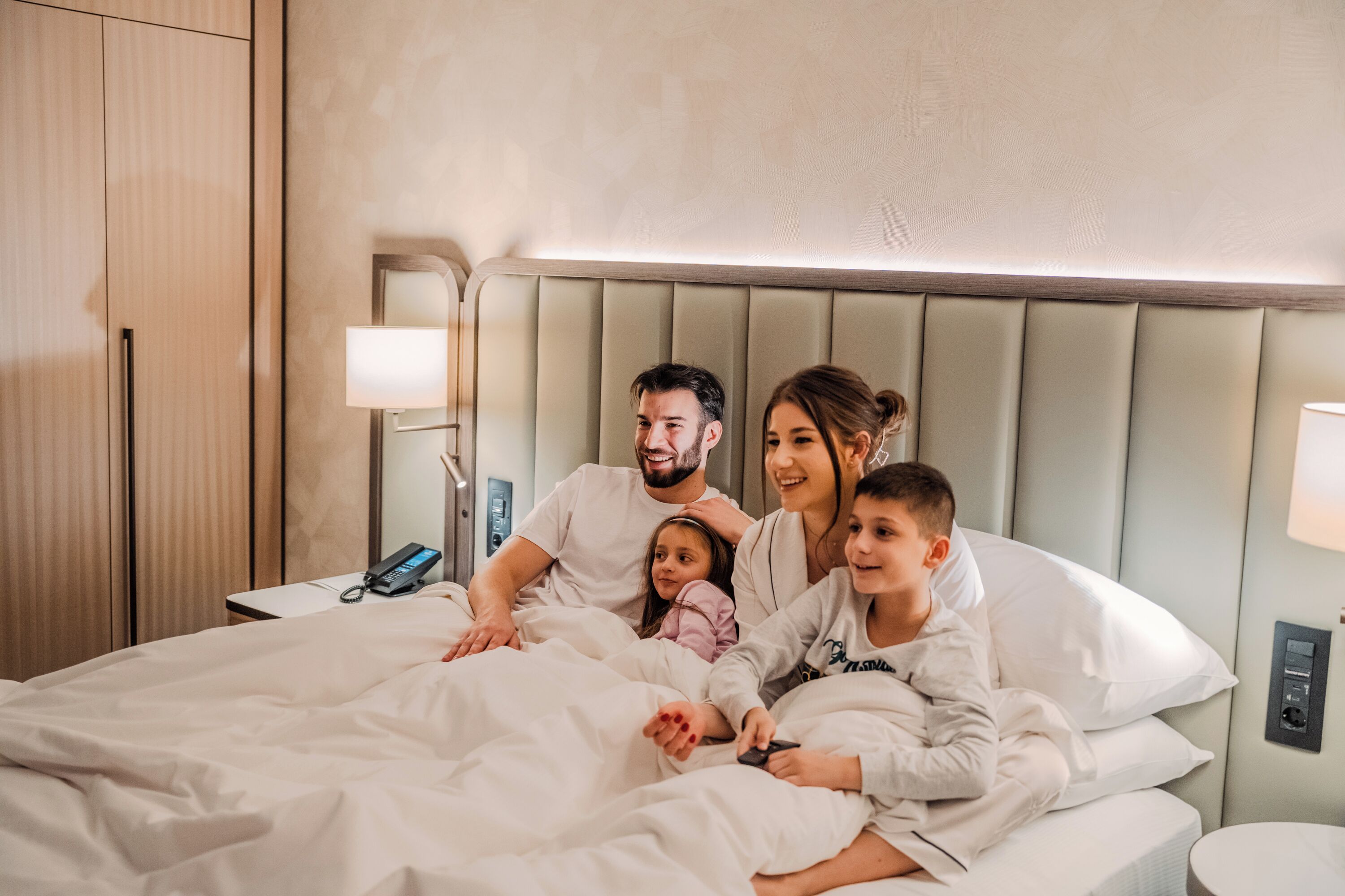 Una familia de cuatro personas en una habitación de hotel con una cama, mesitas de noche y un teléfono en la pared.
