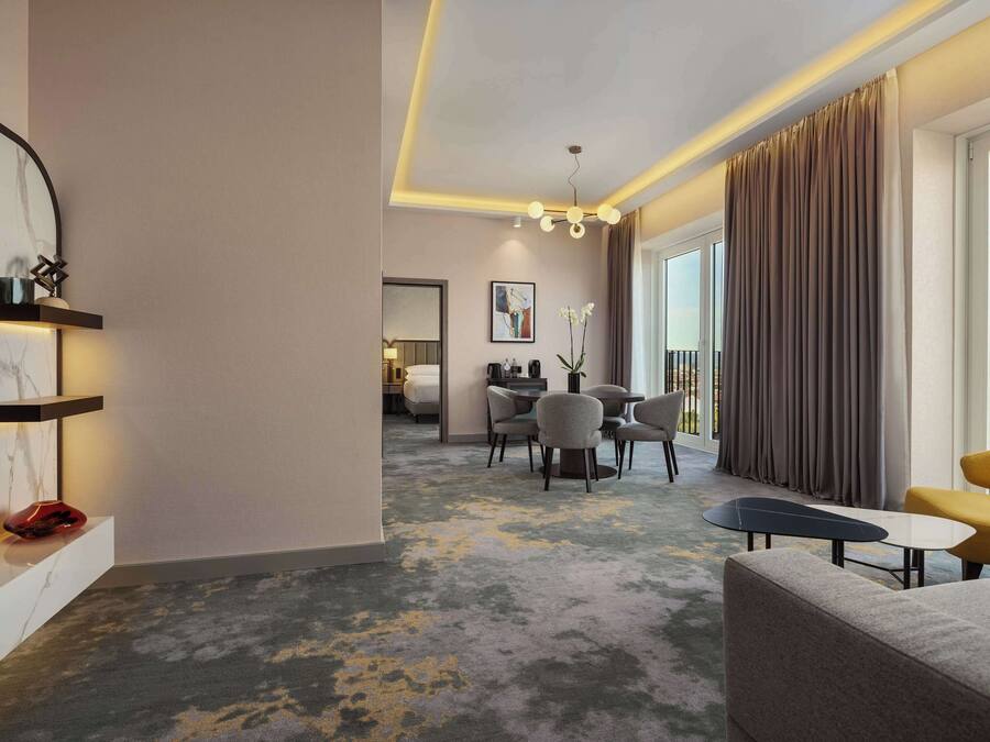 Una habitación de hotel moderna con zona de estar, comedor y balcón con cortinas.