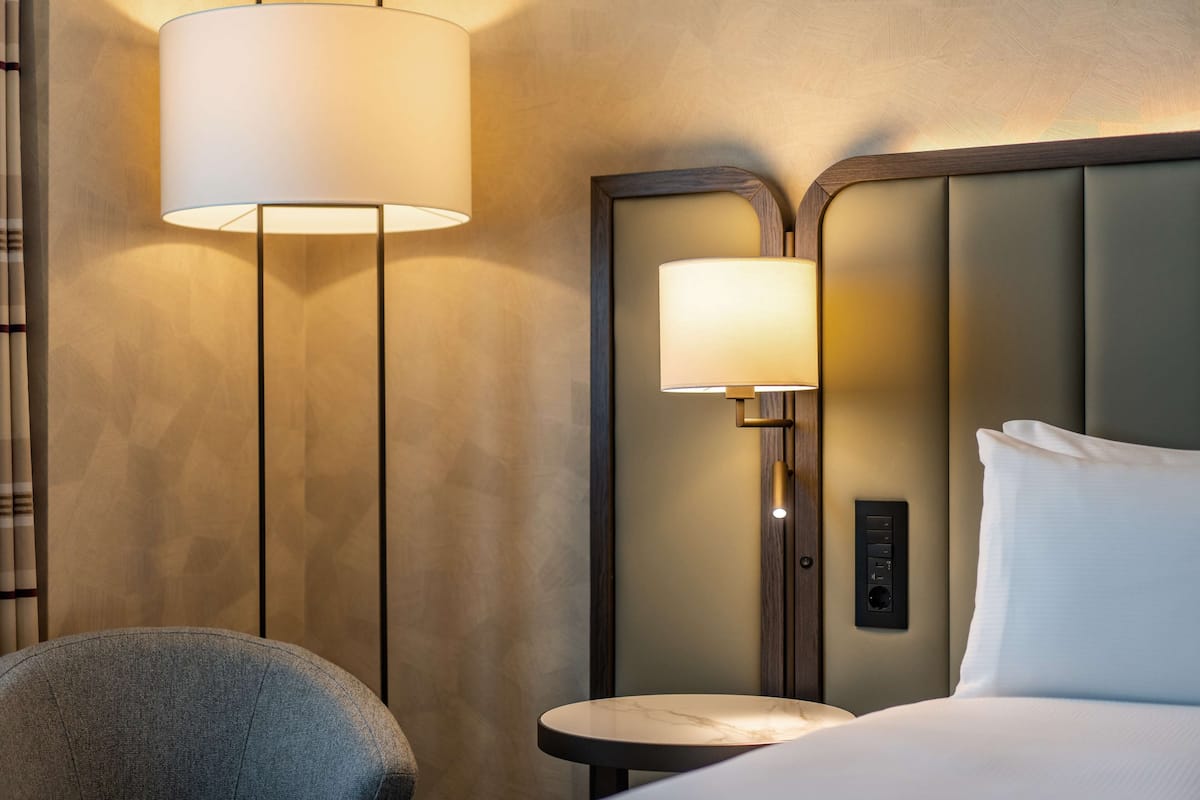 A hotel room with a bed, a round side table, a chair, and two wall-mounted lamps.