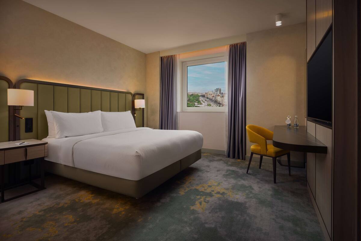 A hotel room with a bed, a desk with a lamp, a chair, a television, and a window with curtains.