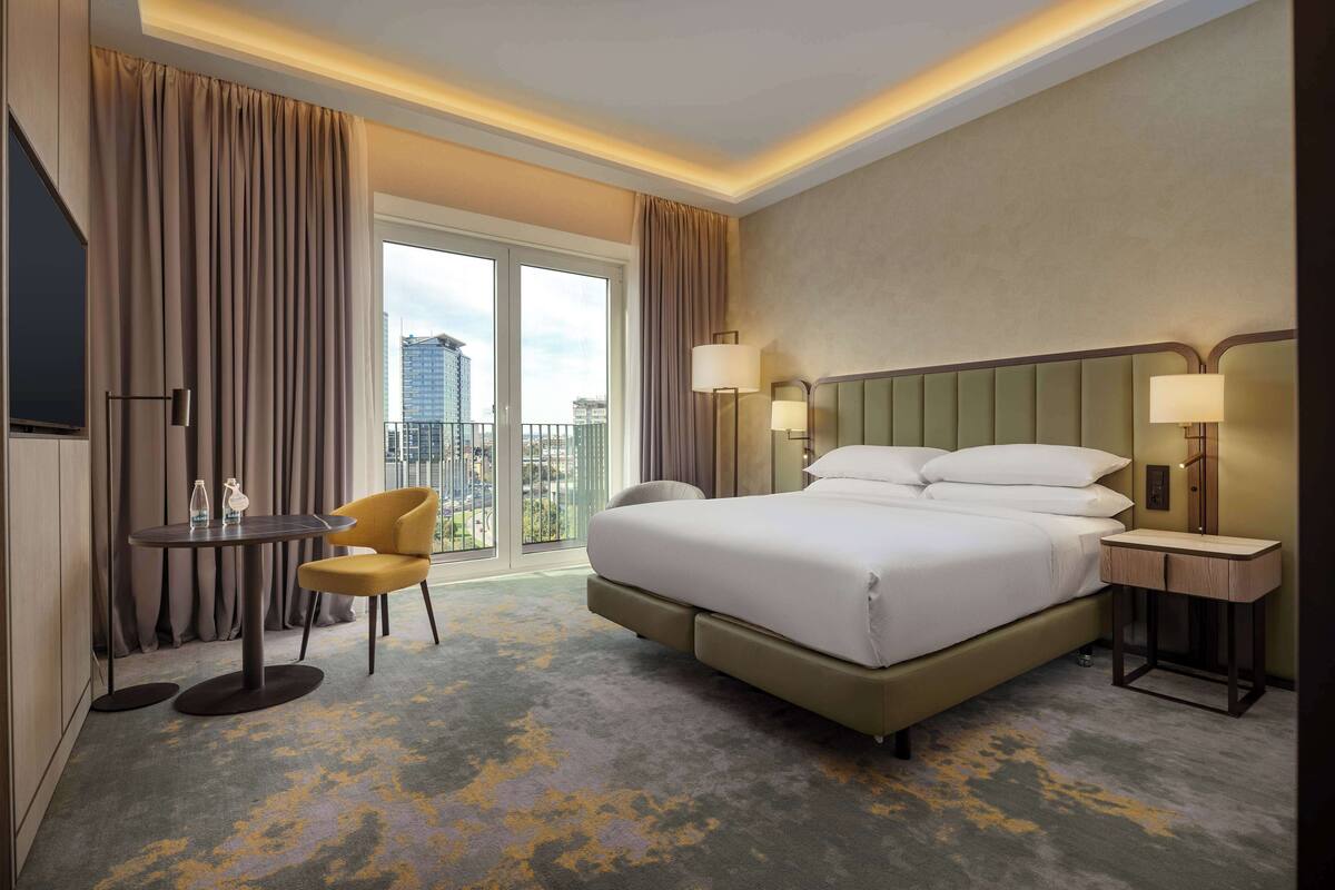 A hotel room with a large bed, a desk with a chair, a lamp, and a view of the city through the window.