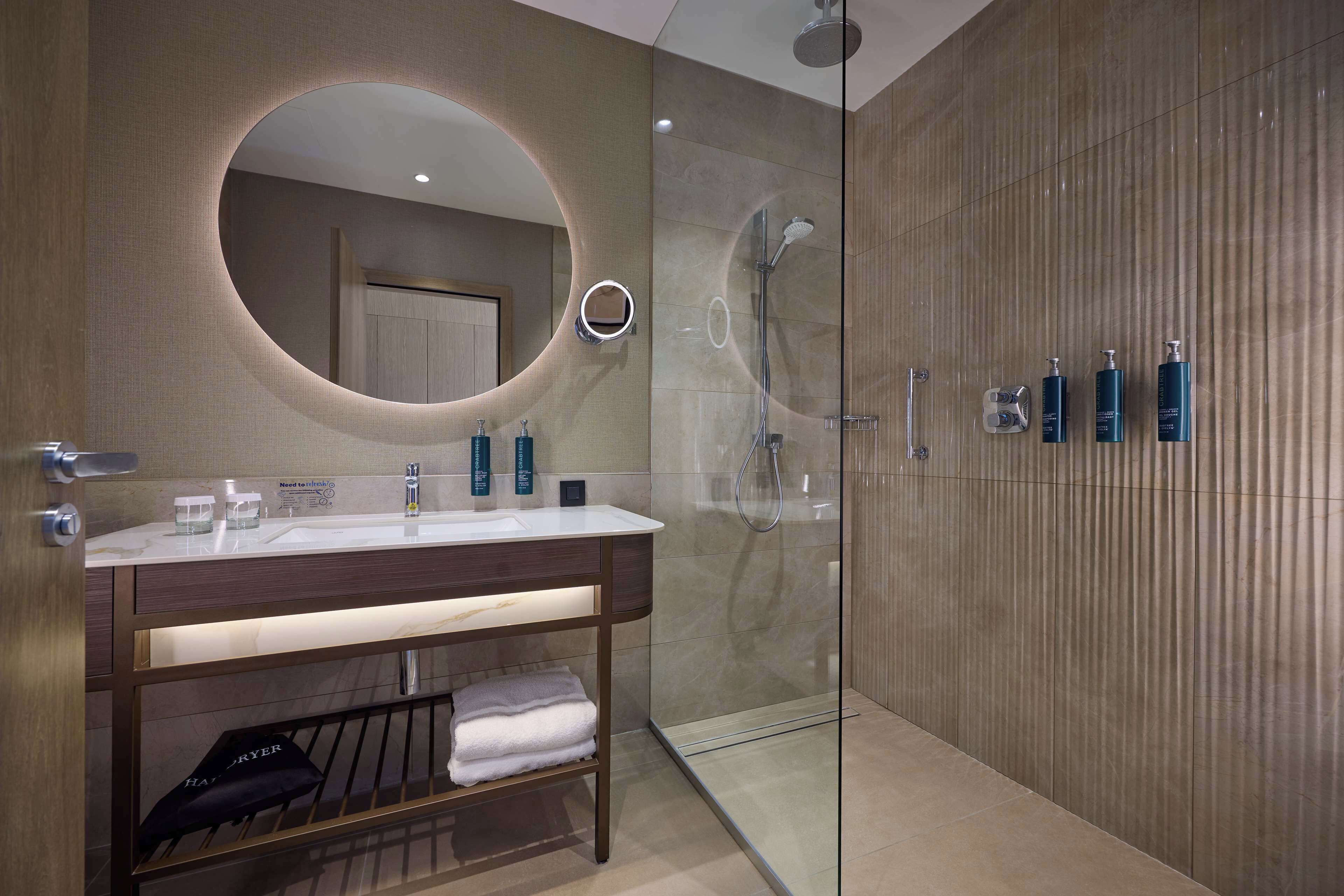 Un baño moderno con un espejo redondo, una ducha con cerramiento de vidrio y artículos de tocador ordenados.