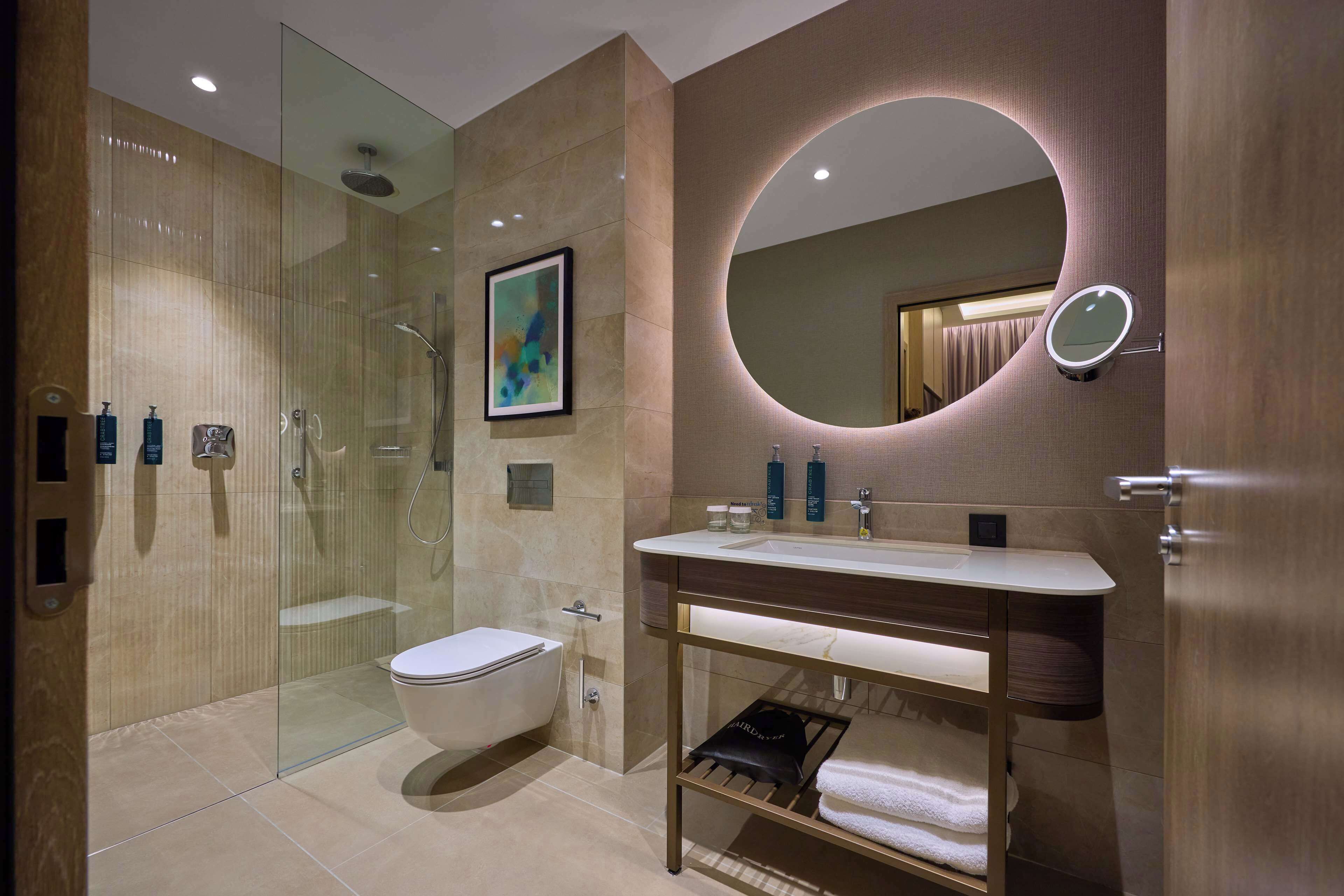 A modern bathroom with a glass-enclosed shower, a toilet, a sink with a vanity, and a mirror with a circular light.