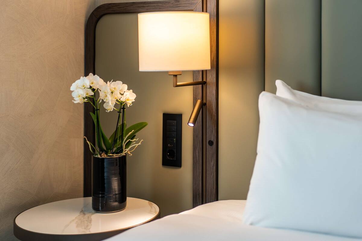 A hotel room with a bed, a bedside table with a vase of white flowers, a wall-mounted lamp, and a control panel.