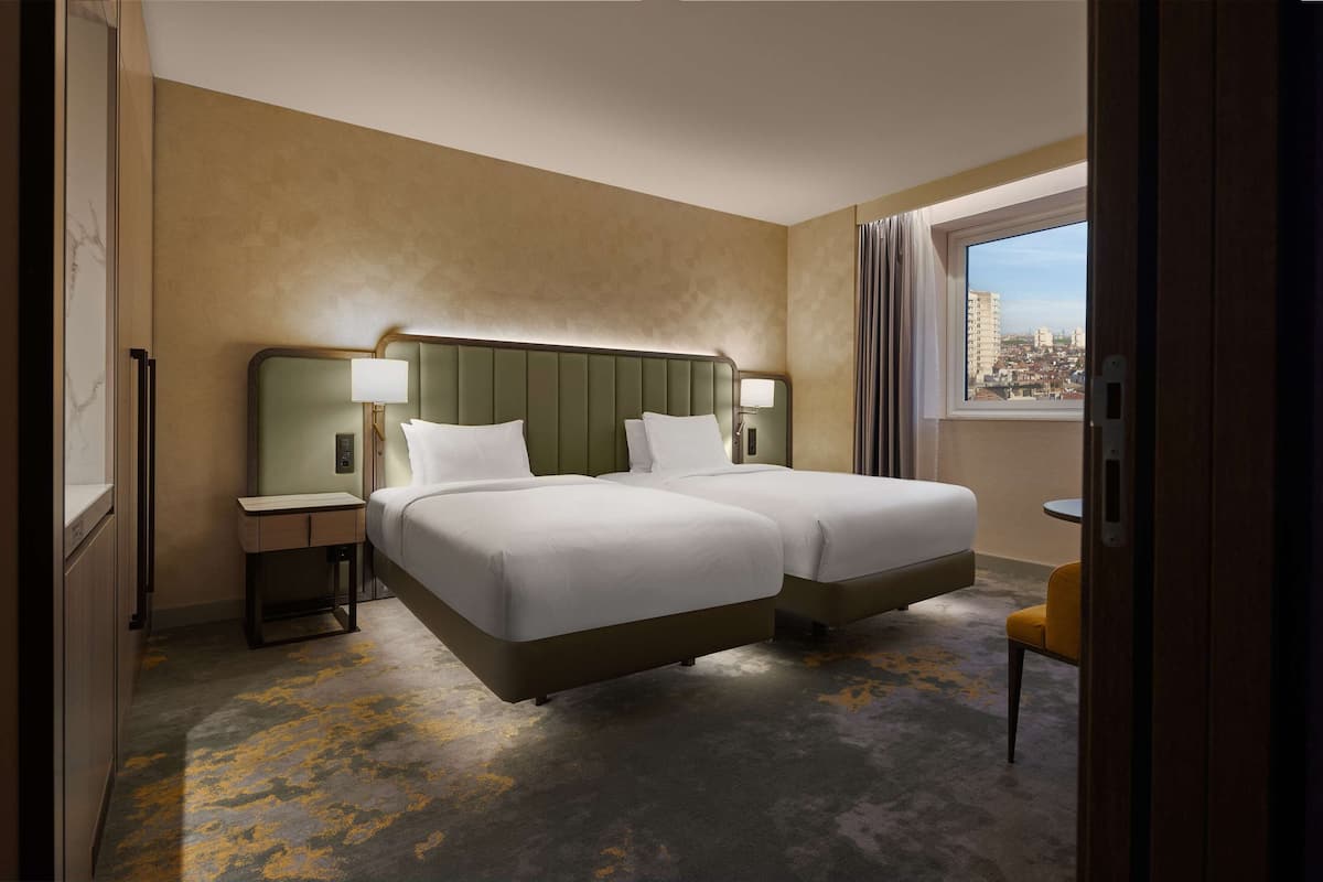 A hotel room with two beds, a small table, a chair, and a window with a city view.