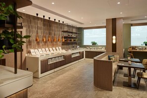 A modern cafe with a central counter displaying various items, surrounded by seating areas and a large window providing a view. - Hilton Sofia (Sofia)