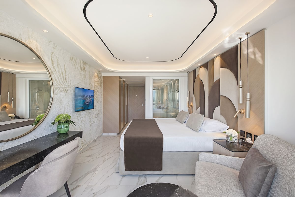 A modern hotel room with a large bed, a sitting area with a sofa and a small table, a large mirror, and a view of the city through the window.