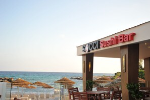 A seaside restaurant with a 'Sushi Bar' sign, outdoor seating with umbrellas, and a view of the ocean.