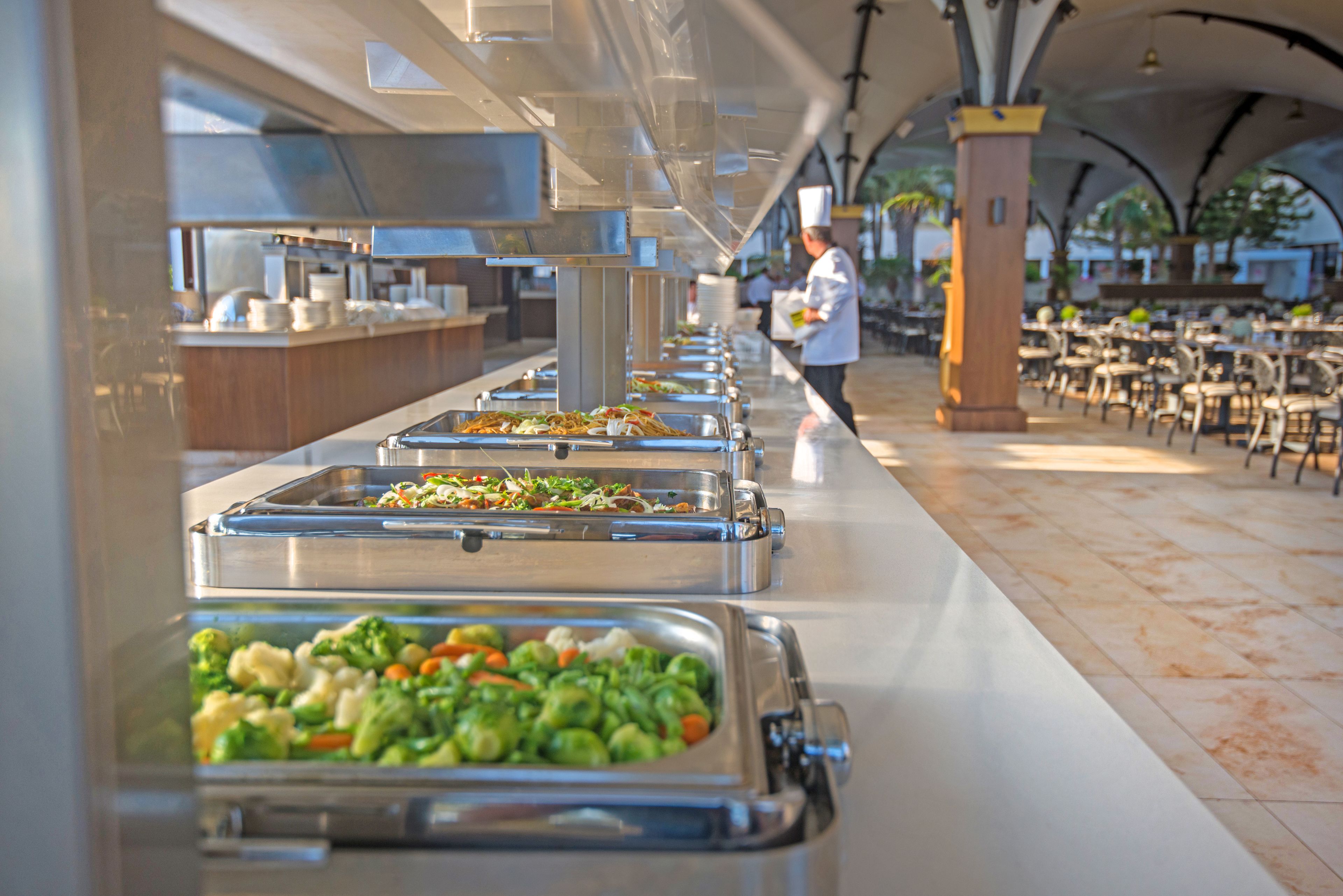 Disposición de bufé con diferentes alimentos en bandejas, un chef al fondo y una zona de estar con mesas y sillas.