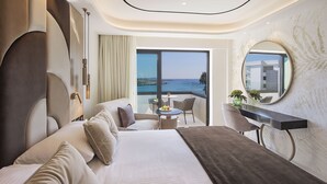 A modern hotel room with a large bed, a sitting area with a table and chairs, a balcony with a view of the sea, and a round mirror on the wall. - Adams Beach Hotel & Spa (Ayia Napa)