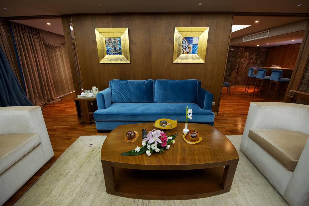 A modern living room with a blue sofa, a wooden coffee table, and a white armchair. There are two framed artworks on the wall and a small table with a vase of flowers.