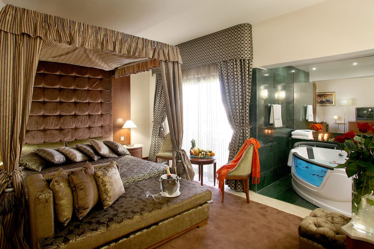 A bedroom with a four-poster bed, a sitting area with a small table, and a bathroom with a bathtub and a mirror.
