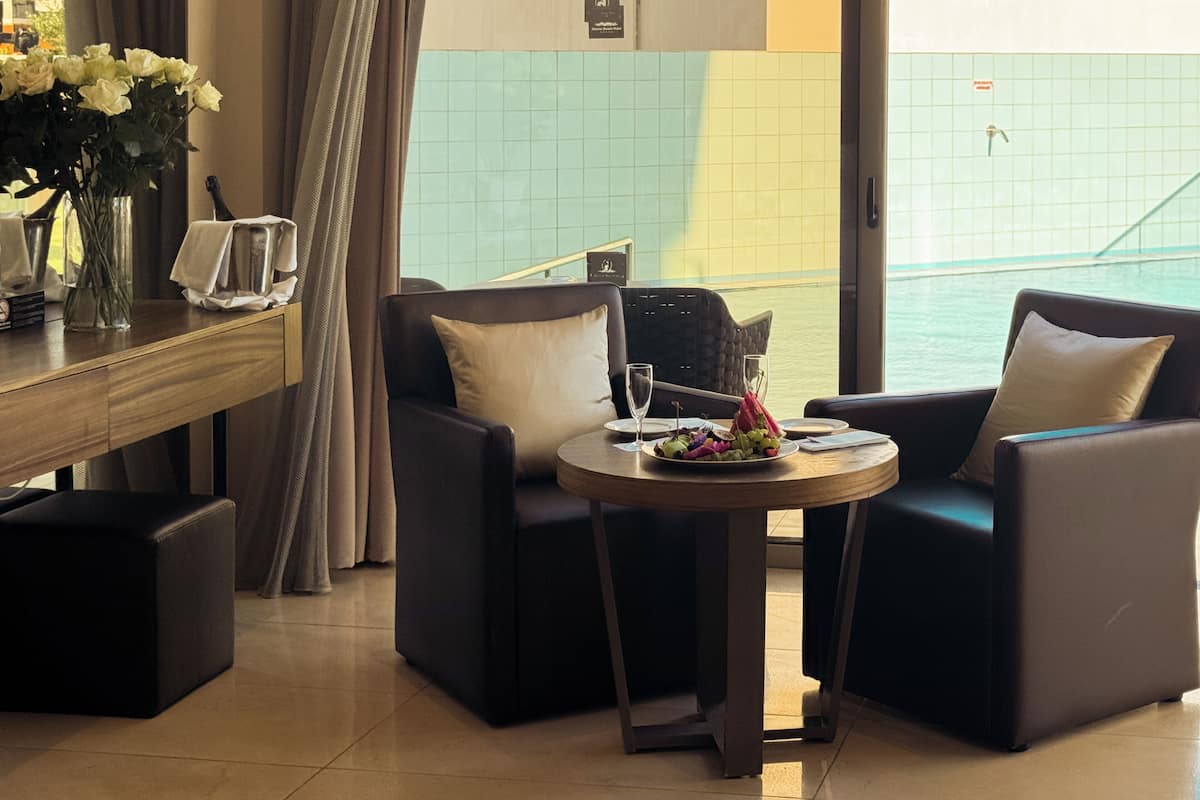 A hotel room with a dining area featuring two chairs, a table with a plate of food and glasses, a vase with flowers, and a view of the sea through a window.