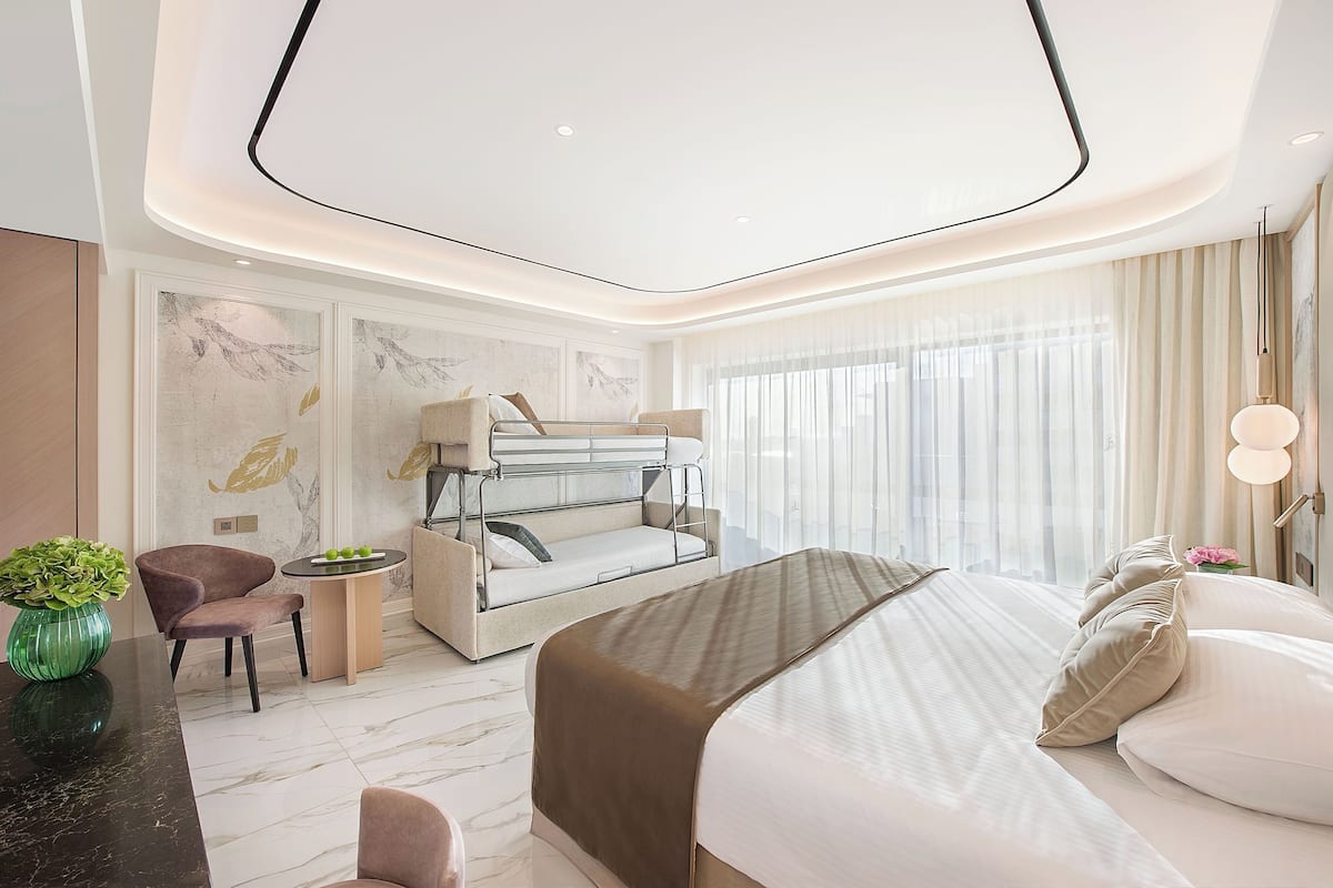 A modern hotel room with a bunk bed, a sitting area with a chair and table, a vase of flowers, and a large window with sheer curtains.