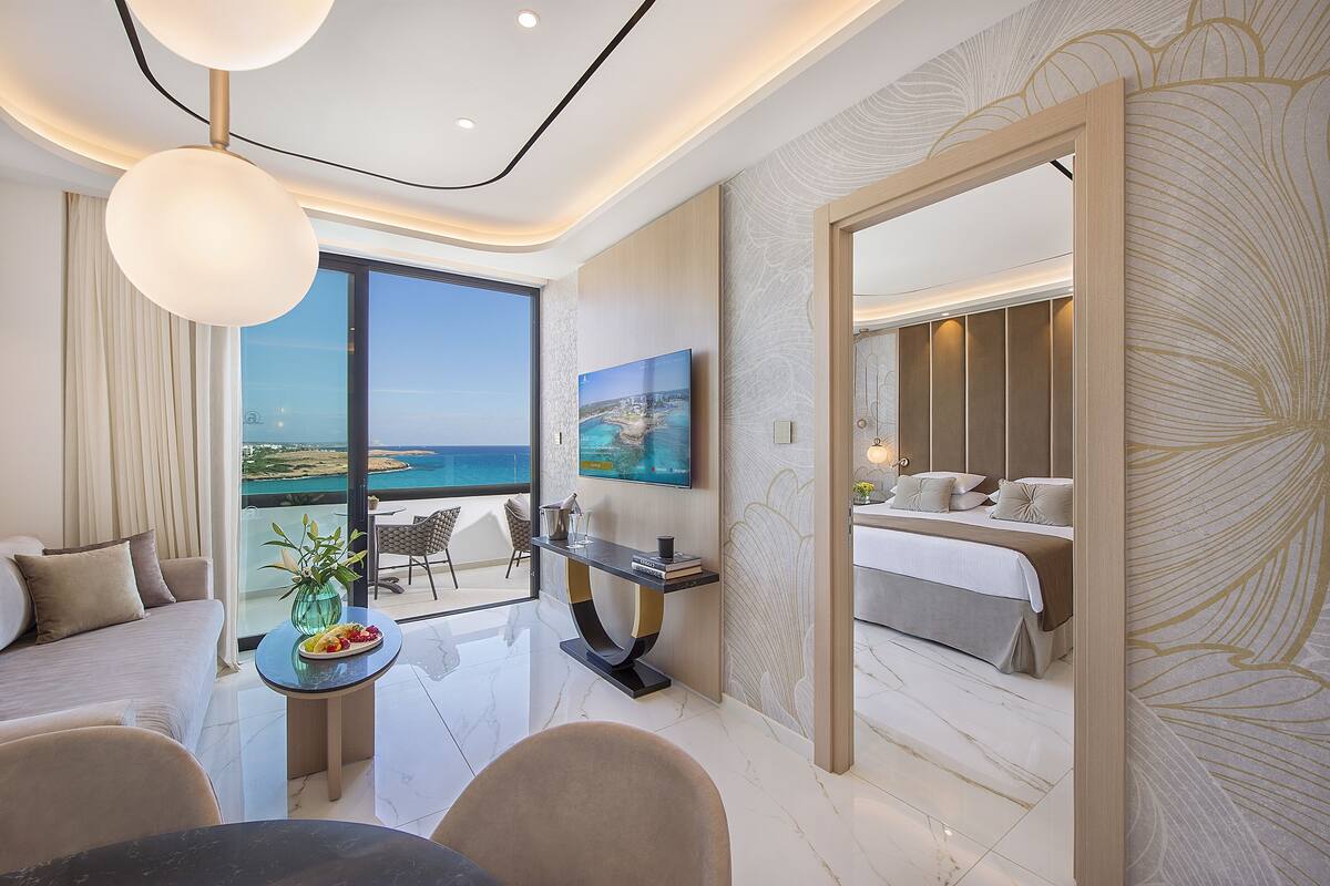 A modern hotel room with a balcony, a large TV, a dining area, and a bedroom with a bed and nightstands.