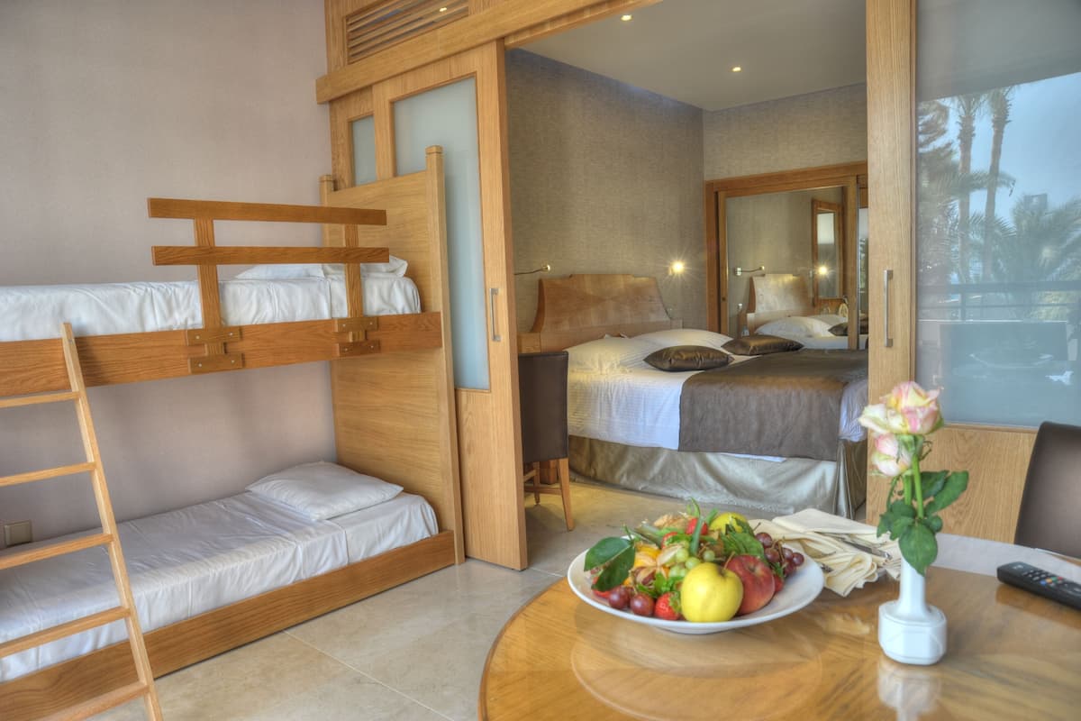 A modern hotel room with a bunk bed, a single bed, a dining table with fruit and a vase, and a balcony with a view of palm trees.