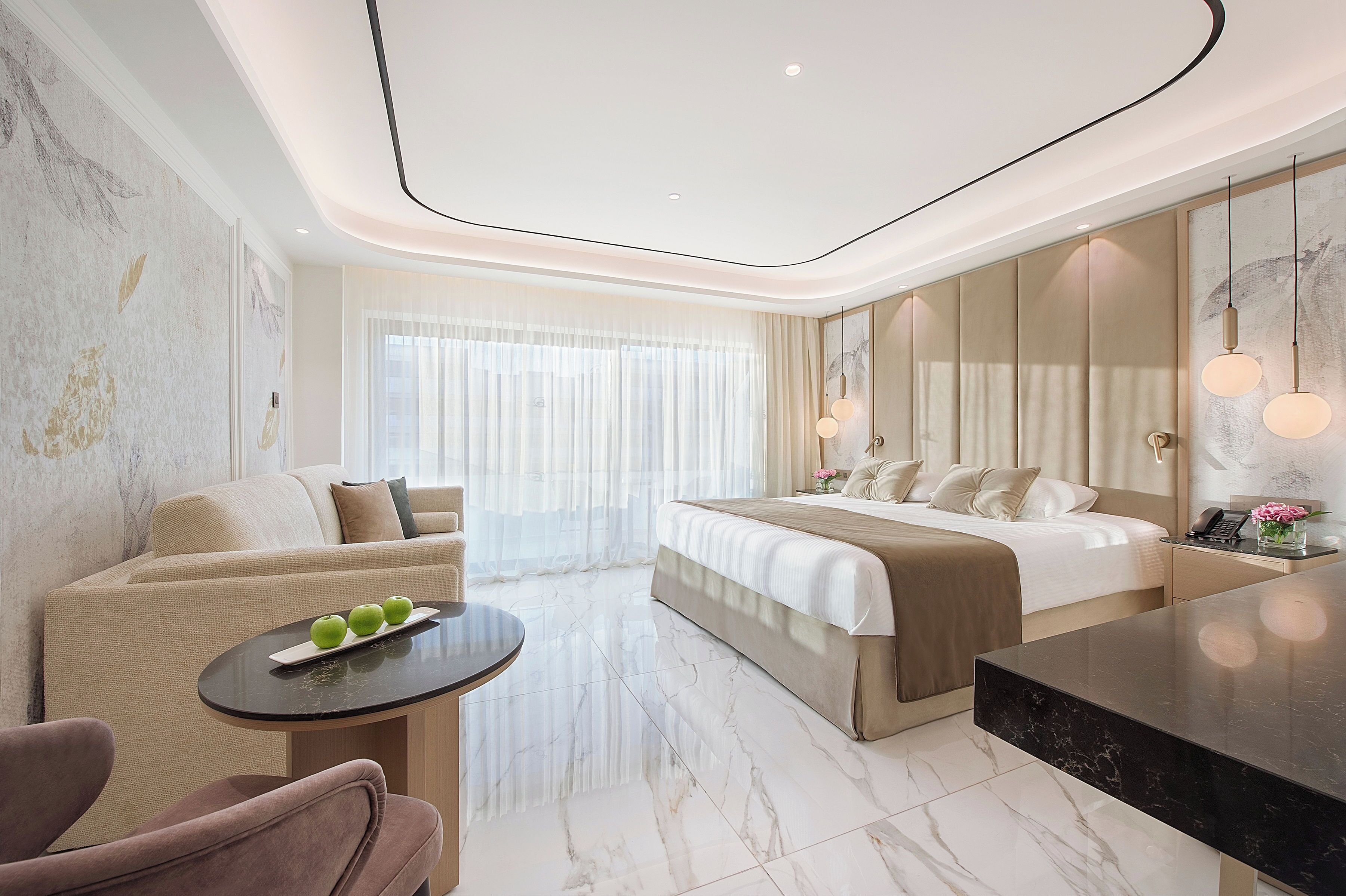 A modern hotel room with a bed, a sitting area with a table, and a large window with sheer curtains.