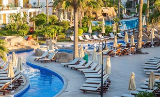 A resort pool area with lounge chairs and umbrellas, surrounded by palm trees and a rocky landscape.