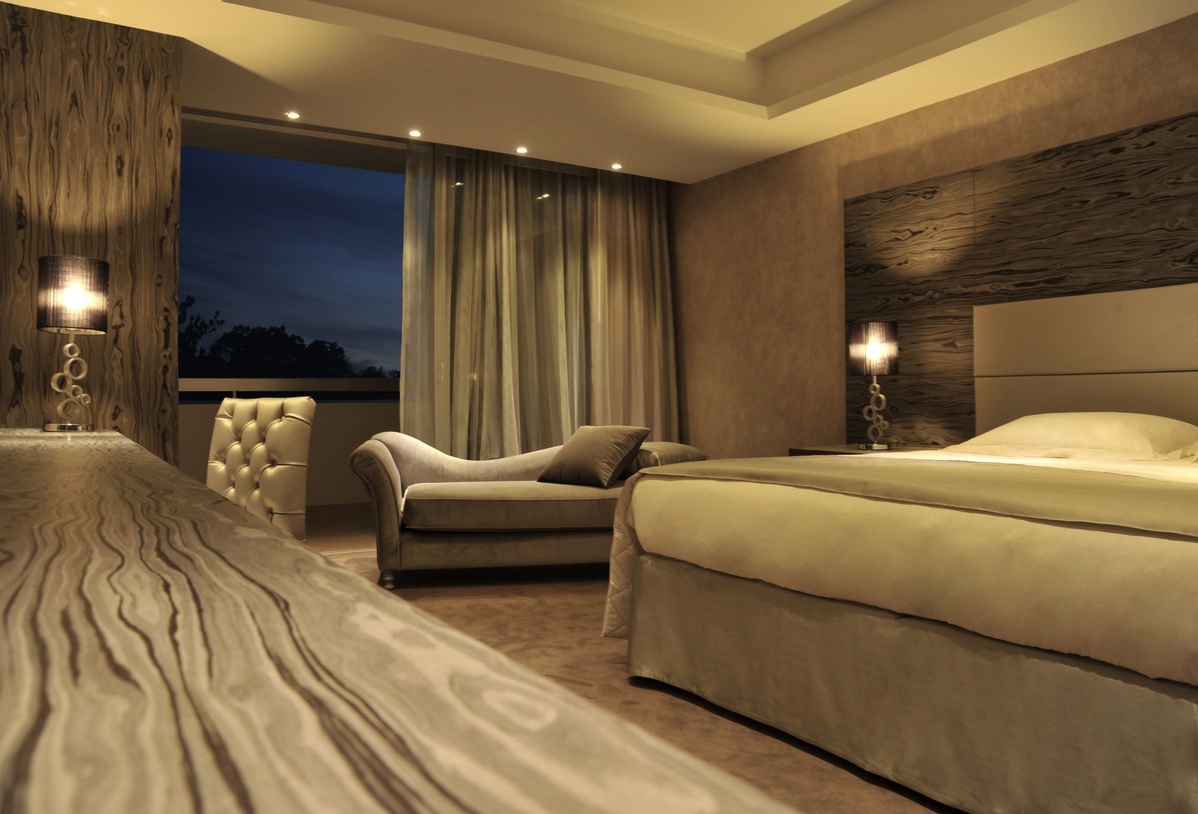 A modern bedroom with a large bed, a sofa, and a chair. There is a view of the outdoors through a large window.