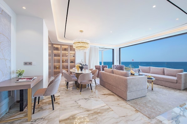 A modern living room with a marble floor, a round dining table with chairs, a sofa, and a large window overlooking the sea.