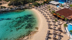 An aerial view of a beach resort with clear turquoise waters, a sandy beach, and numerous sun loungers and umbrellas. - Adams Beach Hotel & Spa (Ayia Napa)