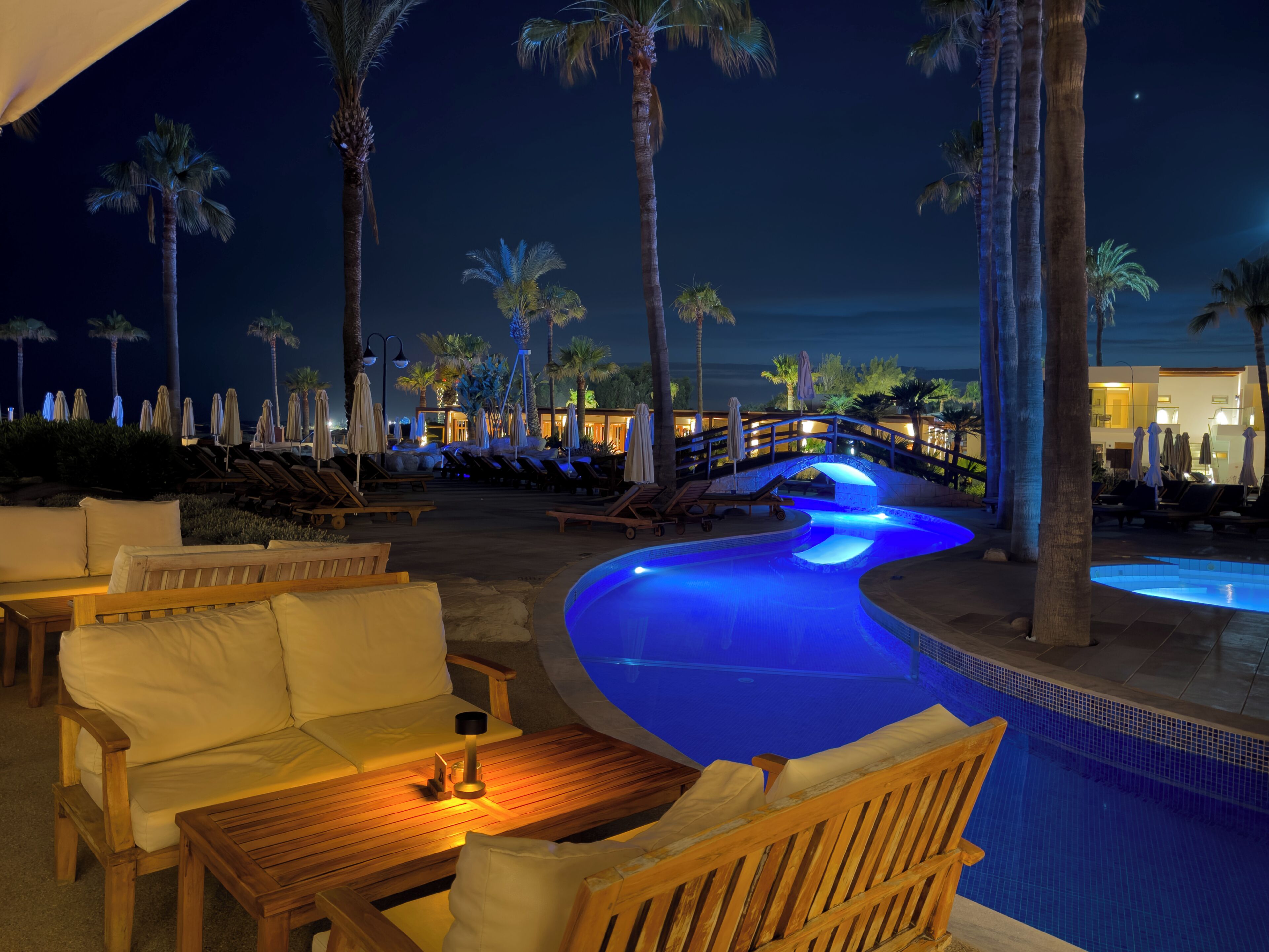 Zona de piscina nocturna con sillones de descanso, una mesa de madera y un puente.