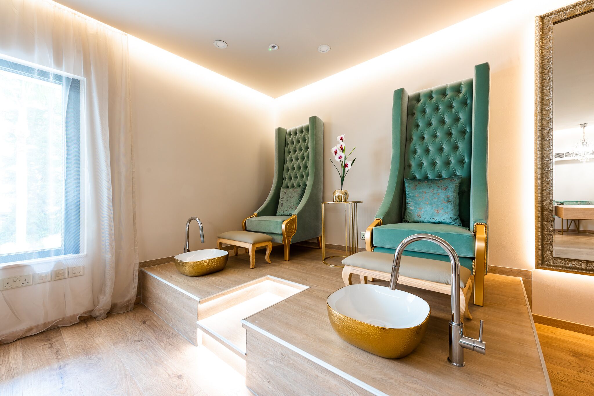 Un baño moderno con un espejo grande, dos sillones verdes acolchados, un lavabo dorado y un suelo de tonos claros.