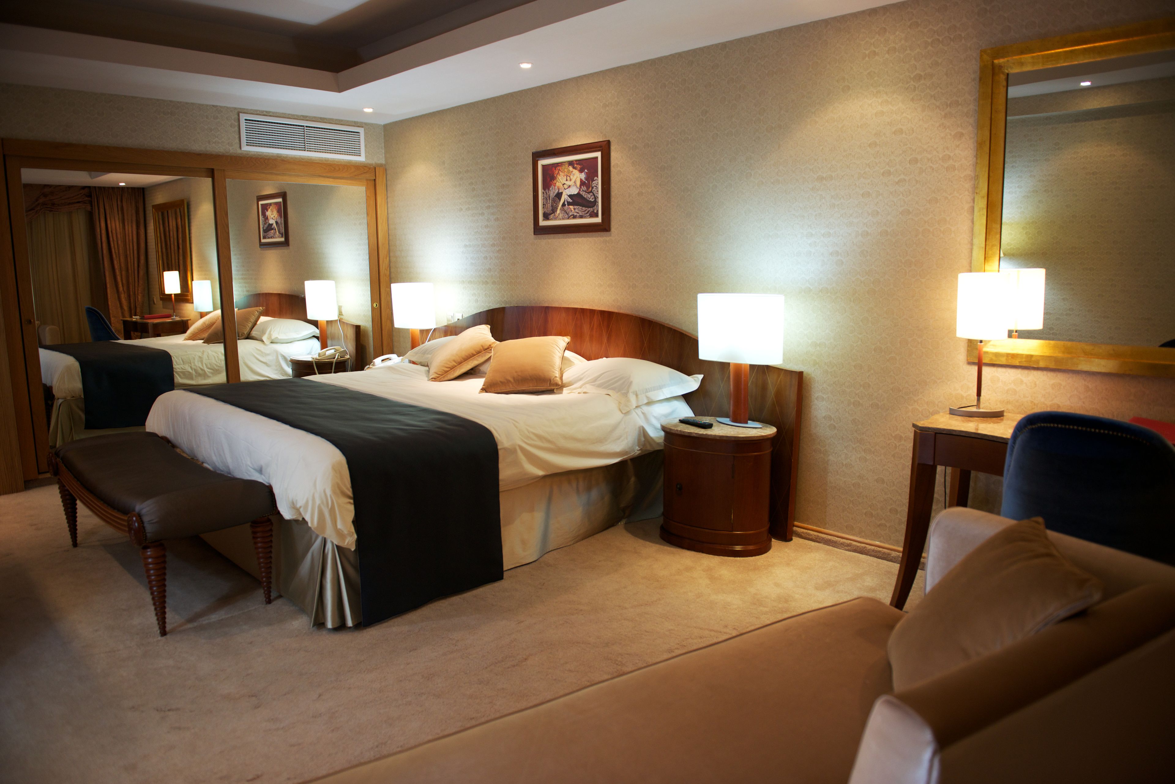 Une chambre d’hôtel avec un grand lit, deux tables de chevet avec des lampes, un banc, une chaise et un grand miroir au mur.