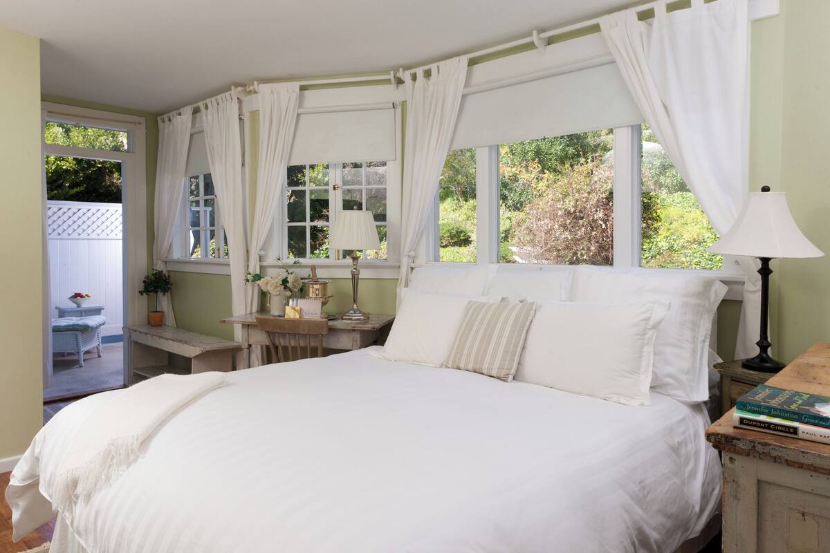 A bedroom with a large bed, a nightstand with a lamp, a window with curtains, and a view of greenery outside.